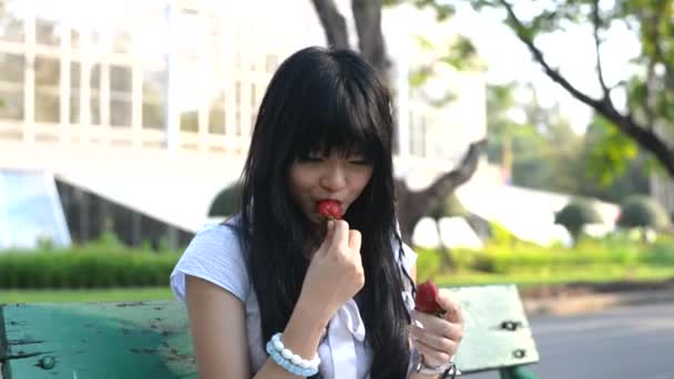 Una linda chica tailandesa asiática con vestido de ropa vintage está sentada en un banco al aire libre comiendo fruta de la comida de fresa en el banco en las vacaciones de verano con expresión facial agria feliz . — Vídeo de stock