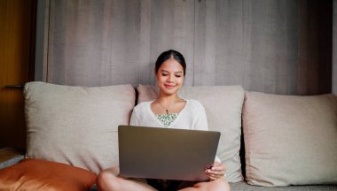 Evdeki oturma odasında dizüstü bilgisayarla çalışan güzel, gülümseyen Asyalı bir kadın. Ev ofisinde çalışan Asyalı iş kadını. Evde eğleniyorum..