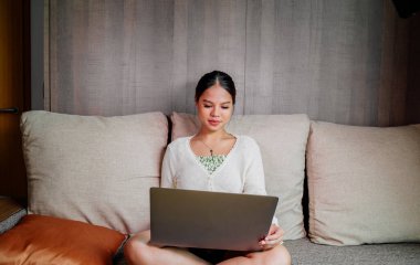 Evdeki oturma odasında dizüstü bilgisayarla çalışan güzel, gülümseyen Asyalı bir kadın. Ev ofisinde çalışan Asyalı iş kadını. Evde eğleniyorum..