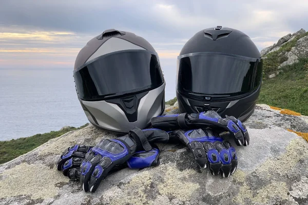 两个摩托车头盔的特写 带着色眼镜和皮革摩托车手套在岩石上 背景美丽的海景 免版税图库图片