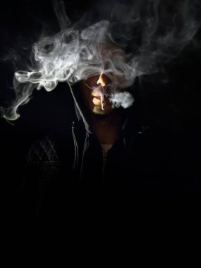 Adam sigara içiyor. Siyah bir kapüşonlu giyiyor. Sigara yanıyor ve onu ağzında tutuyor.