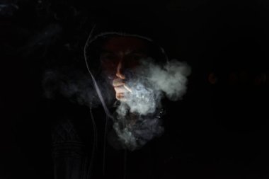 Bir adam sigara içiyor. Siyah bir kapüşonlu giyiyor. Sigara yanıyor ve onu ağzında tutuyor.