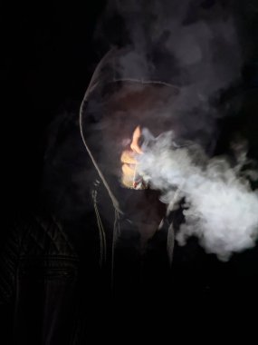 Adam sigara içiyor. Siyah bir kapüşonlu giyiyor. Sigara yanıyor ve onu ağzında tutuyor.