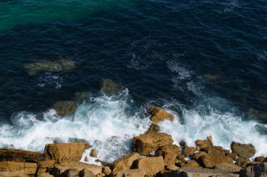 Kayalık okyanus kıyısındaki sörf çizgisi, arka plan, su ve kahverengi kayaların koyu mavi rengi.