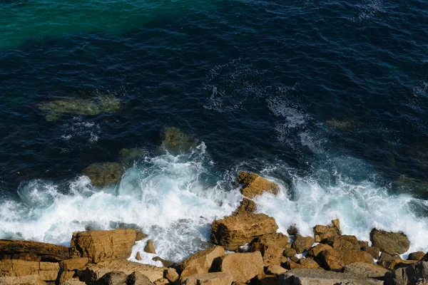 Kayalık okyanus kıyısındaki sörf çizgisi, arka plan, su ve kahverengi kayaların koyu mavi rengi.