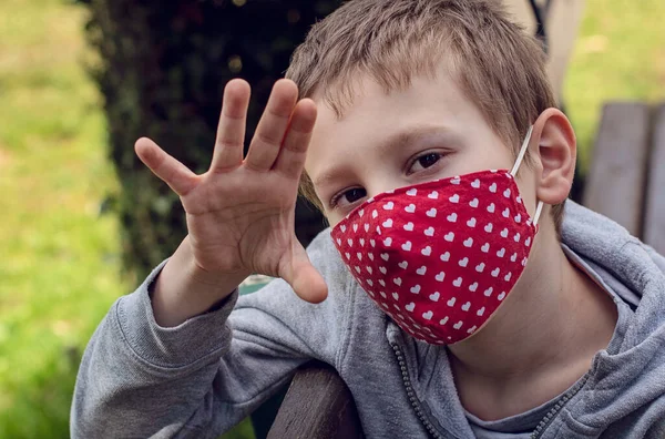 Portrait of young boy wearing handmade cotton face mask. Boy in mask in the park in sunny day. Protection against saliva, cough, dust, pollution, virus, bacteria, coronavirus, COVID-19.