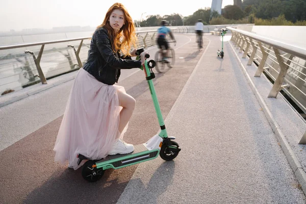 Red haired pretty girl using electric scooter in fashinable long pink dress black leather jacket. sunny Morning in metropolis big city. alternative eco-conscious transport.  bicycle riders way to work