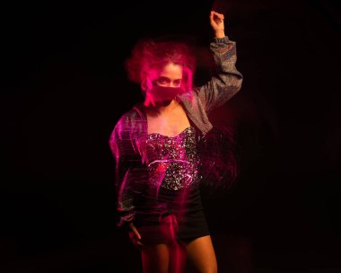 Parlak, şenlikli, siyah arka planlı ve koruyucu, hijyen maskeli güzel bir genç kadın. Uzun pozlama sanatsal yaratıcı dinamik hareket efekti. Kaldırılmış el gergin endişe dolu görünüyor.