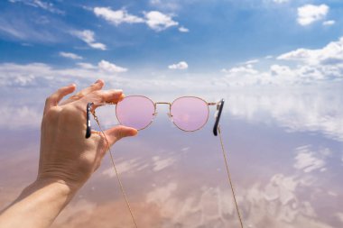 Pembe gölün önünde pembe gözlüklü bir kadın. Güneşli güzel bir gün. Bulutlar ve mavi gökyüzü. Tatil zamanı mutluluk havası. Yazın tadını çıkar. Hipster blog yazarı tarzı. Tatil afişi planlanıyor