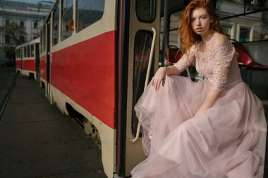 Doğu Avrupa toplu taşımacılığı. Kızıl saçlı kızıl saçlı harika genç kız eski Sovyet tramvayının merdivenlerinde oturuyor. Kalitesiz moda balo tarzı. Pembe retro elbise. Sert ayakkabılar. 