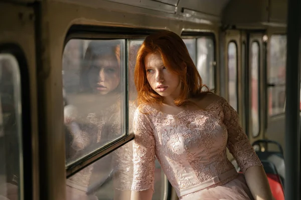 dramatic portrait of a girl near the window in an old Soviet tram. Beautiful attractive red haired girl looking out the window. Retro pink lace fancy dress. sad brooding nostalgia emotion. youth rebel