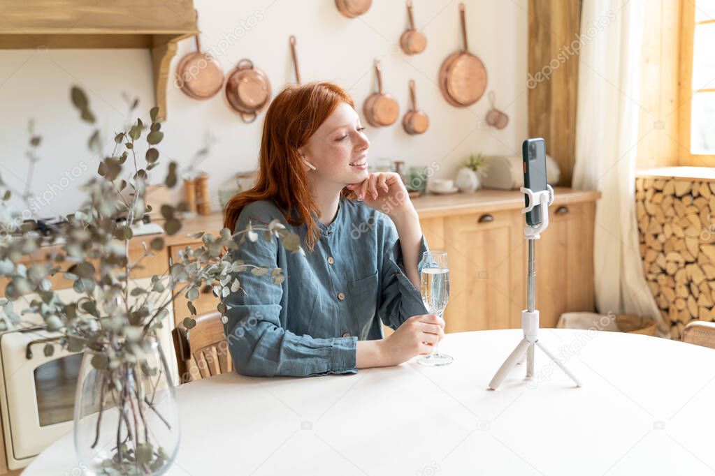 Smiling video call. long distance relationship remote celebration online. Girl at kitchen holding glass of champagne using cell phone online video call conversation. Blue linen short. Lockdown time