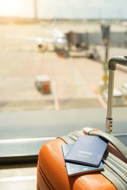 Pandemik zaman dikey fotoğrafı sırasında havaalanı. Üzerinde aşı pasaportu olan turuncu bir çanta. Uçak arka planda kalkışa hazırlanıyor. Hava alanında güneşli bir yolculuk günü. Sağlıklı güvenlik turisti