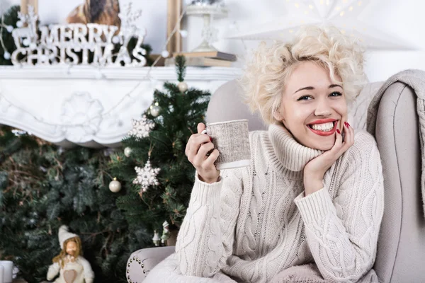 Bella grande sorriso donna bionda seduta nel comfort accogliente della sedia a casa con una coperta che indossa una maglia maglione maglia — Foto Stock