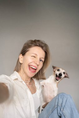 Havalı, flört eden sarışın kadın ve geniş mutlu gülüşlü komik küçük köpek. Stüdyoda gri arkaplanlı selfie. Evcil arkadaşınla iyi vakit geçir. boş kopyalama alanı
