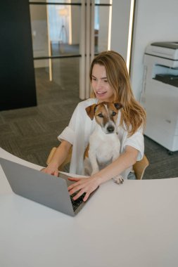 Birlikte çalışan mutlu arkadaşlar. Kadın ve evcil hayvan tedavisi Jack Russell Terrier ofiste dizüstü bilgisayarla çalışıyorlar. yaratıcı açık alan. Sarışın genç köpek ebeveyn kadın. dikey kompozisyon. Köpek kameraya bakıyor. duygusal destek