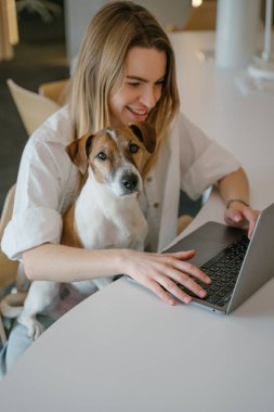 Mutlu kadın ve evcil hayvan tedavisi Jack Russell Terrier birlikte çalışma alanında dizüstü bilgisayarla çalışıyorlar. Yaratıcı ofis açık alan. Sarışın genç köpek ebeveyn kadın. dikey kompozisyon. Gülümseyen genç köpek sahibi.