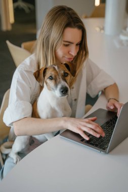 Sarışın kadın ve evcil hayvanı Jack Russell Terrier çalışma alanında dizüstü bilgisayarla birlikte çalışıyorlar. Yaratıcı ofis açık alan. Sarışın genç köpek ebeveyn kadın. dikey kompozisyon. Köpek kameraya bakıyor.