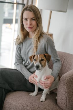 Evcil hayvanla seyahat. Gülümseyen sarışın kadın ve köpek Jack Russell Terrier pembe sandalyede oturmuş kameraya bakıyor. Evcil hayvana sarılan bir kadın. Doğal ışık. dikey bileşim