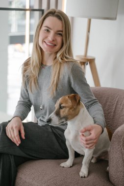 Gülen Sarışın kadın ve köpek Jack Russell teriyeri pembe sandalyede oturmuş kamera köpeğine bakıyor. Doğal ışık. dikey bileşim