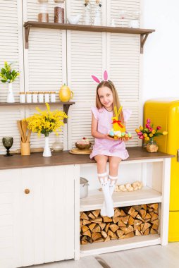 Mutlu Paskalyalar. Çocuk Paskalya için hazırlanıyor. Tavşan kulaklı bir kız süslü bir mutfakta oturur ve Paskalya yumurtalarını tutar...