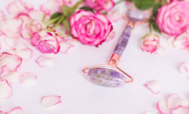 The front roller is made of amethyst on a white background with pink roses. Massager for lifting the skin made of natural stone. Close-up, selective focus.