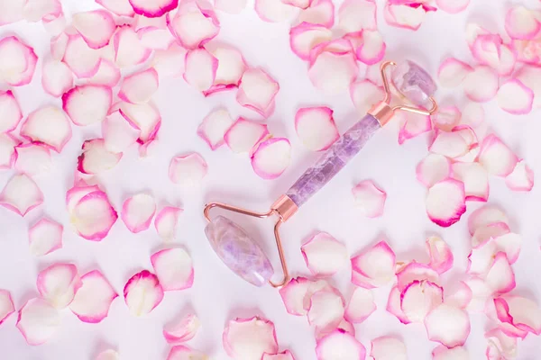 Massager, face roller. Roller made of natural amethyst stone on a white background with pink rose petals..