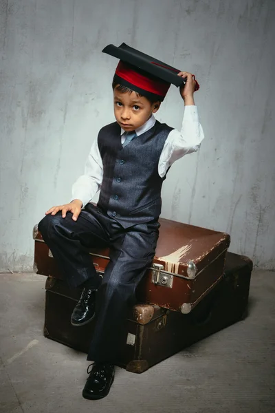 small black boy in  graduation hat sits on the old suitcases