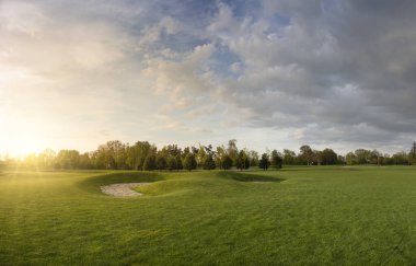 Çimenli golf sahası. Parkta akşam manzarası. Bulutlu mavi gökyüzü ve gün batımında ışıldayan parlak güneş ışığı