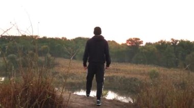 Sporcu kenara yaklaşıyor, göle bakıyor, kırsal kesimdeki ağaçlara bakıyor.