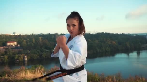 Jovem mulher kimono branco demonstra sua técnica de karatê contra a natureza pano de fundo — Vídeo de Stock