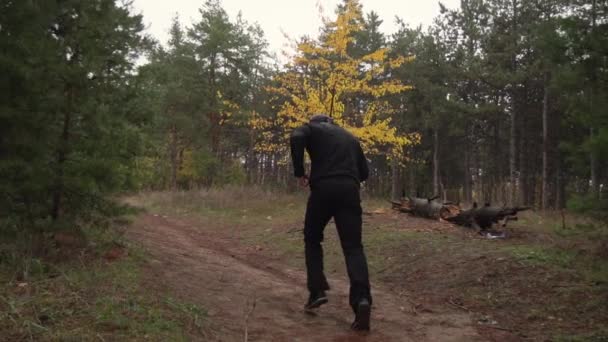 Un tipo con un vestido negro está entrenando, corre por el bosque entre los pinos, otoño — Vídeo de stock