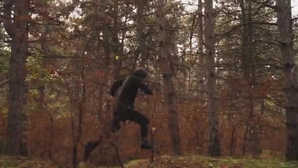 Un type en vêtements noirs court à travers la forêt entre les arbres, sautant par-dessus les billes — Video