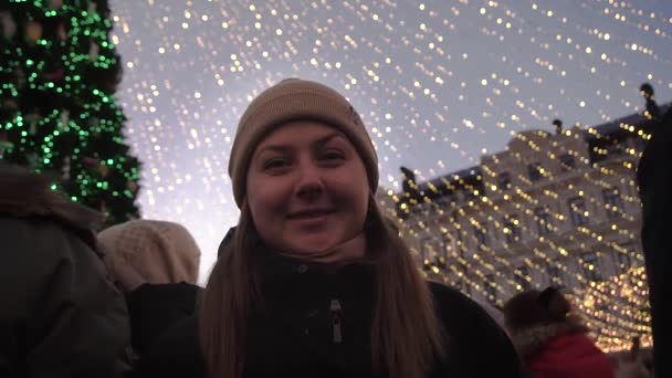 Uma menina sorri para a árvore da cidade, na praça principal no inverno em um chapéu — Vídeo de Stock