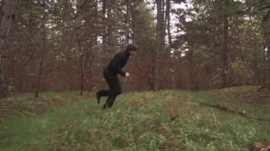 Siyah elbiseli bir adam ağaçların arasında ormanda koşuyor, kütüklerin üzerinden atlıyor.