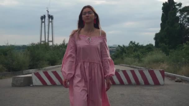 Güzel, gözlüklü kız, pembe elbise kameraya gidiyor. Arkaplanda şehir — Stok video