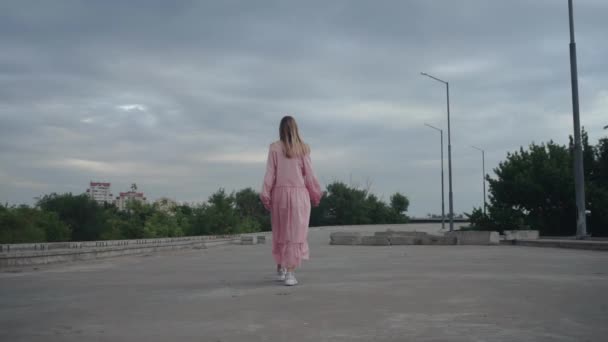 Egy lány sétál egy elhagyatott betonúton, egy befejezetlen hídon. — Stock videók