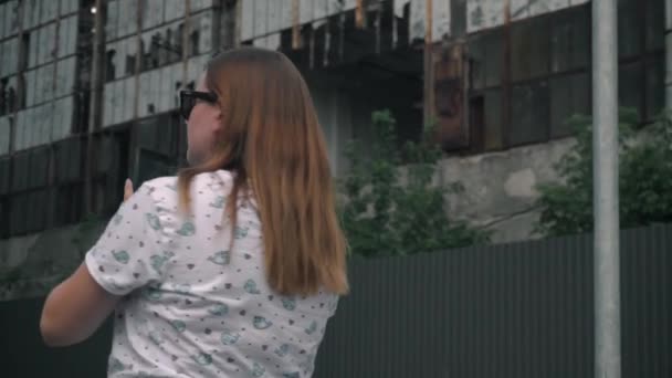 Mooi meisje met een zonnebril kijkt naar de telefoon, dan rond. op de achtergrond een verwoest gebouw — Stockvideo