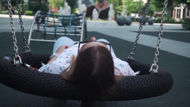 Flicka med mörkt hår rullar långsamt på en gunga, avslappnad, vila. framför en lekplats, fontäner — Stockvideo