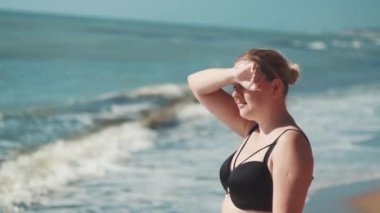 Mayolu bir kız deniz kıyısında duruyor ve parlak güneş yüzünden yüzünü eliyle kapatıyor.