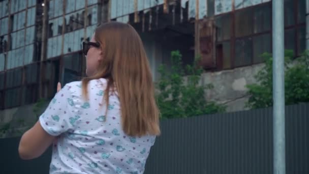 Piękna dziewczyna w okularach przeciwsłonecznych patrzy na telefon, a potem dookoła. w tle zniszczony budynek — Wideo stockowe