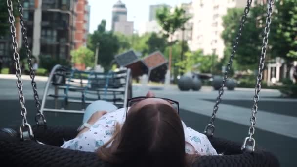 Flicka med mörkt hår långsamt rullande swing, avslappnad, vila. främre lekplats, fontäner — Stockvideo