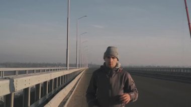 Sıcak giysiler içinde genç bir atlet sabah yürüyüşüne çıktı. Yol köprüsü boyunca, arabalar sürüyor. Sabah sisi arka planda görülebiliyor.