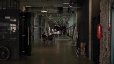 Açık alan modern ofis eski terk edilmiş fabrika, tavanın altında çalışan borular, büyük lambalar asılı, merkez koridor masalar var, asistan işleri dizüstü bilgisayarı, bölüm ofisleri camdan yapılmış.