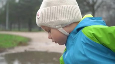Beyaz şapkalı ve mavi yağmurluklu bir çocuk sonbahar yağmurunda bir su birikintisinin yanındaki parkta oynar. Odaklanmış ve özenli, gri bir gün, soğuk.