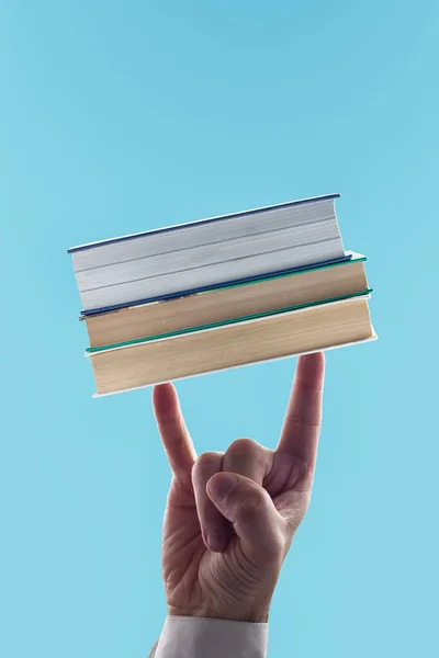 Man\'s hand making rock & roll symbol, holding stack of books. Sign of rock on blue isolated background. Knowledge, wisdom, reading, education and learning concept. Copy space