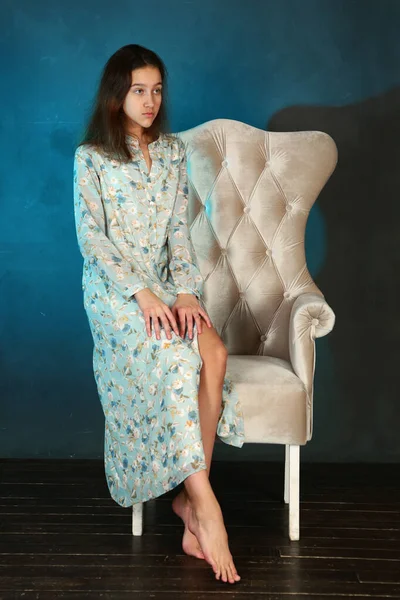 model girl in summer printed dress on vintage armchair and blue wall background full body portrait