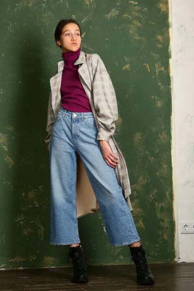 Девушка-подросток в клетчатом плаще, джинсах и ботинках, ходящих по всему телу портрет на фоне зеленой стены — стоковое фото