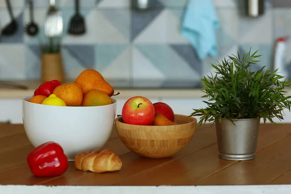 Frutta in ciotola natura morta con pianta in vaso, paprica e croissant sul tavolo foto primo piano su sfondo cucina blu — Foto Stock