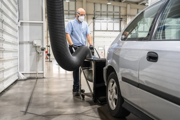 Bir teknisyen, yüz maskesi, eldiven ve gözlüklerle korunan bir aracın periyodik denetiminden önce duman emici yerleştiriyor.